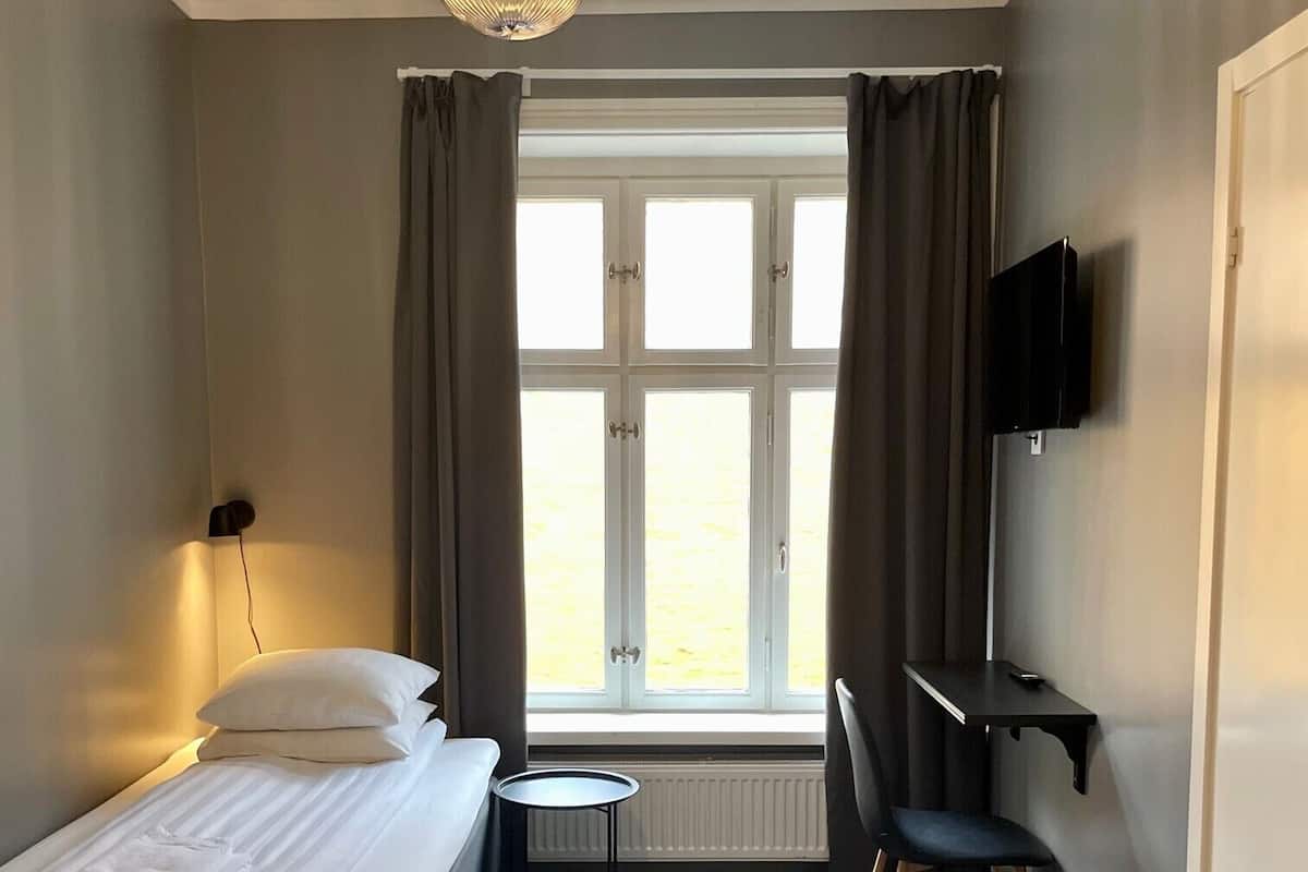 Ein Hotelzimmer mit einem Bett, einem kleinen Tisch, einem Sessel, einem an der Wand montierten Fernseher und einem Fenster mit Vorhängen.