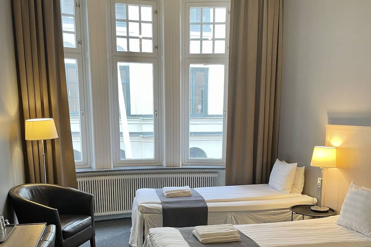 A hotel room with two beds, a desk, a chair, and a lamp.