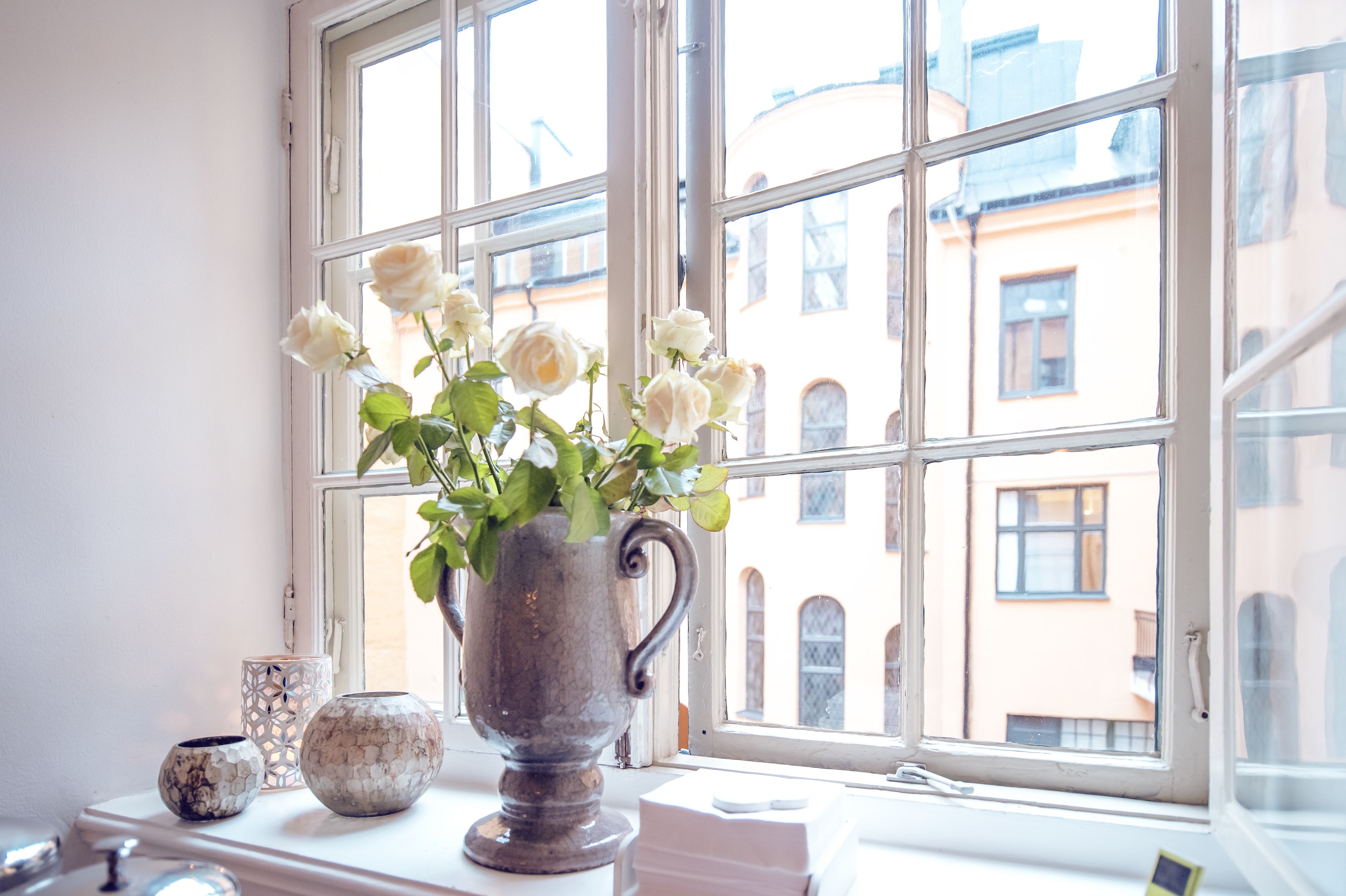 Una ventana con vistas a edificios, un jarrón con flores blancas y dos jarrones decorativos en el alféizar.
