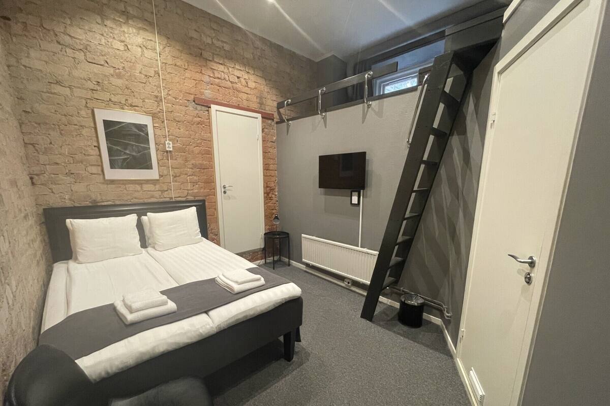 A bedroom with a brick wall, a bed with white bedding, a TV mounted on the wall, and a ladder against the wall.