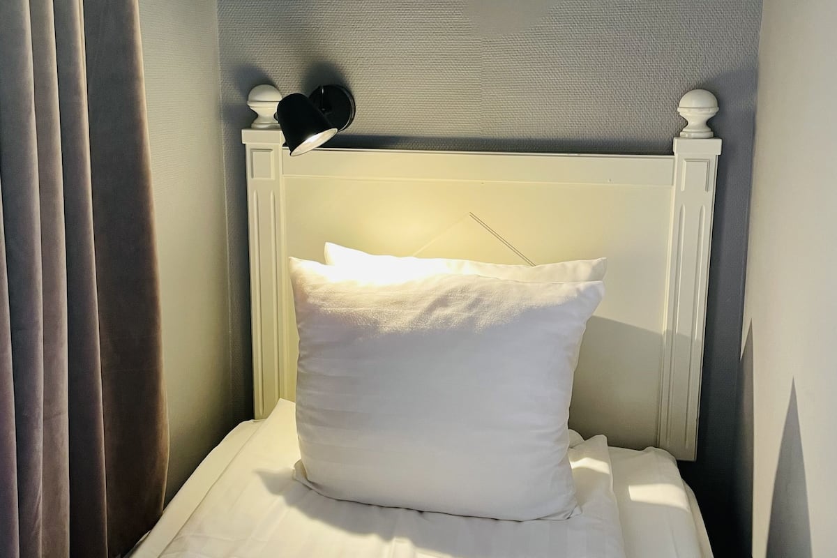 A single bed with white bedding, a headboard with a decorative finial, and a wall-mounted lamp.