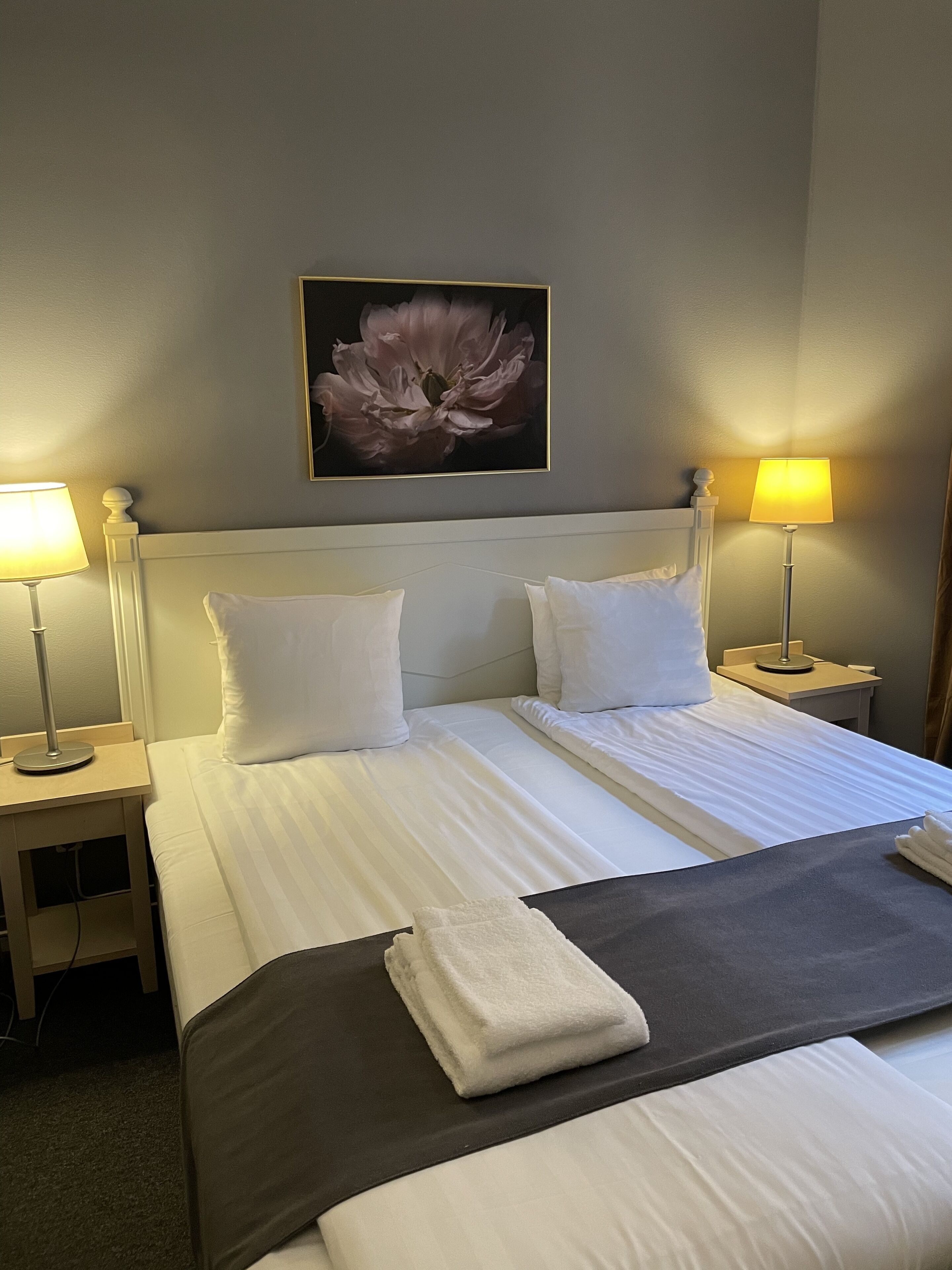 A hotel room with a large bed, two bedside lamps, a wall-mounted flower painting, and a neatly made bed with a folded towel.