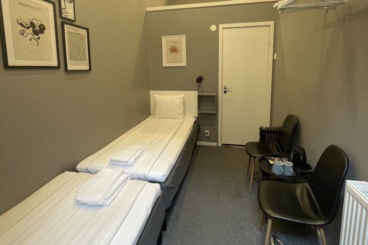 A small, neatly arranged hotel room with two single beds, a desk with a chair, a radiator, and a door leading to another room.