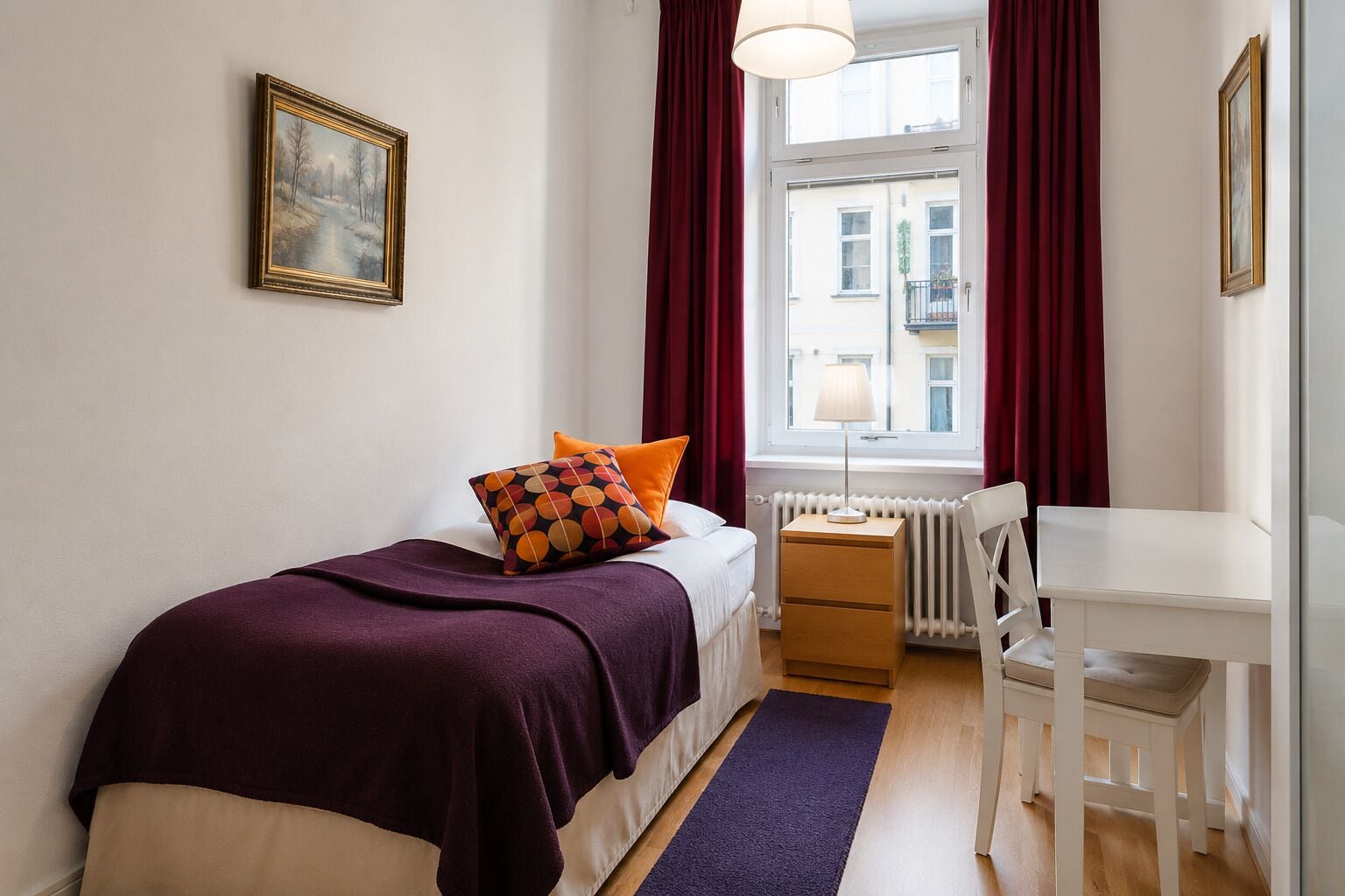 A bedroom with a bed, a desk, a chair, a radiator, and a window with curtains.