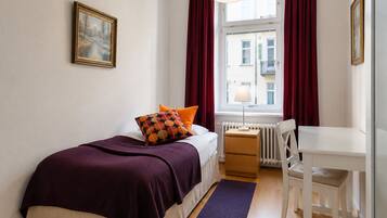 A bedroom with a bed, a desk, a chair, a radiator, and a window with curtains.