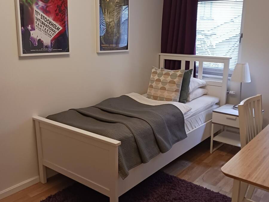 Quarto com cama, escrivaninha, cadeira, janela com cortinas e dois cartazes na parede.