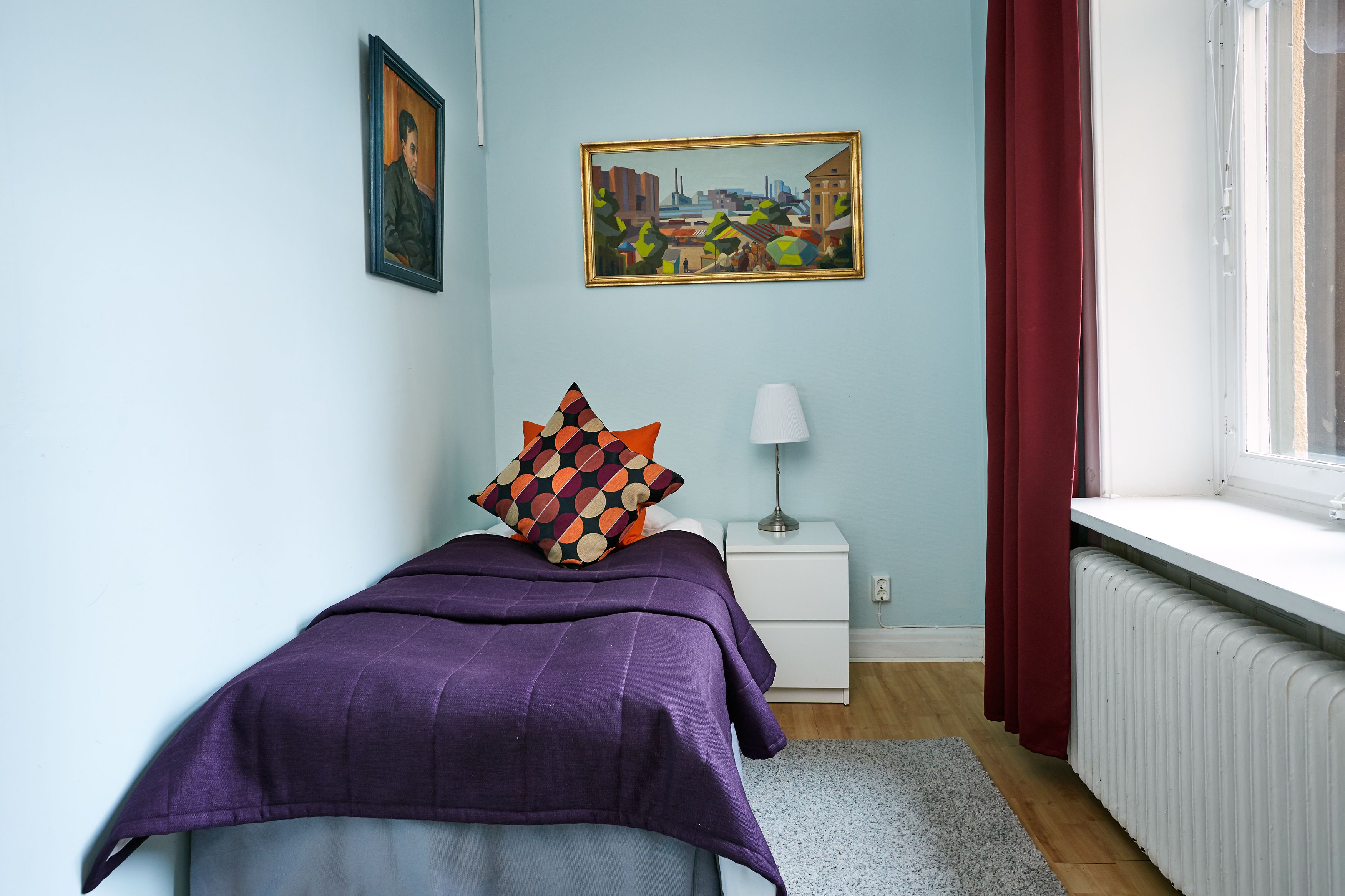 Un dormitorio con cama, mesita de noche, lámpara, ventana con cortinas y un cuadro en la pared.