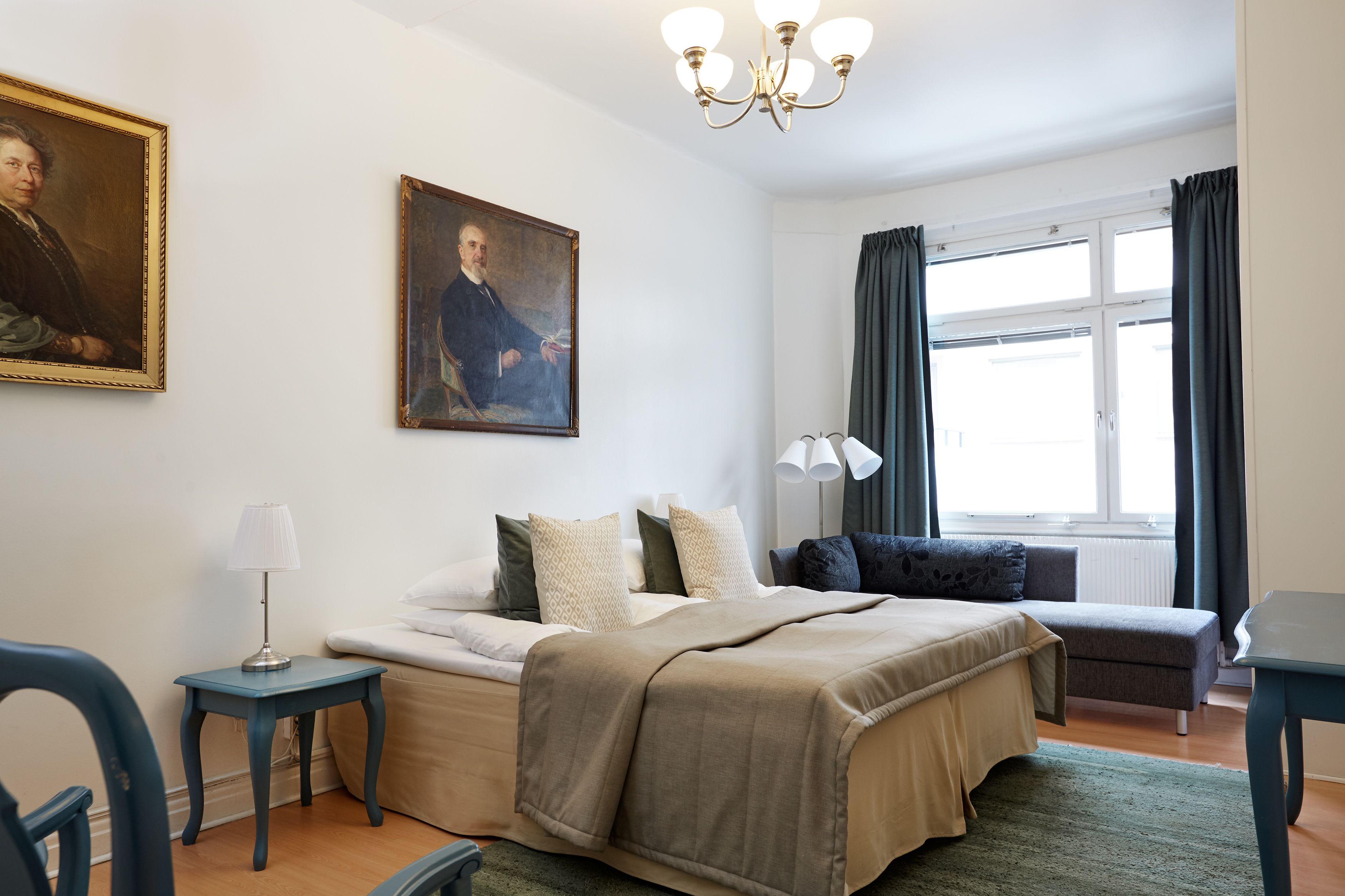 A bedroom with a bed, bedside tables, a desk, a chair, a window with curtains, and a painting on the wall.