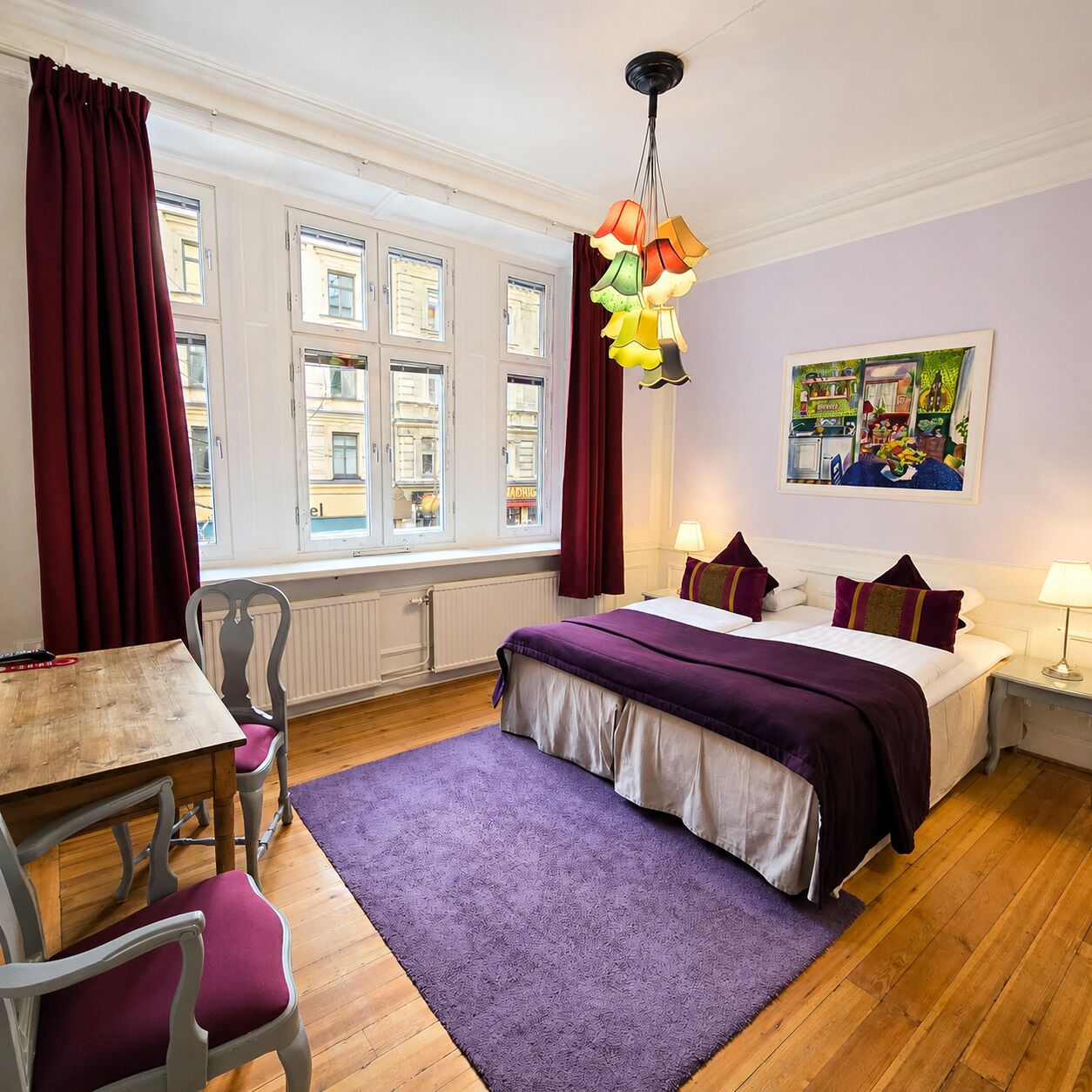 A hotel room with a bed, a desk, a chair, a purple rug, and a window with curtains.