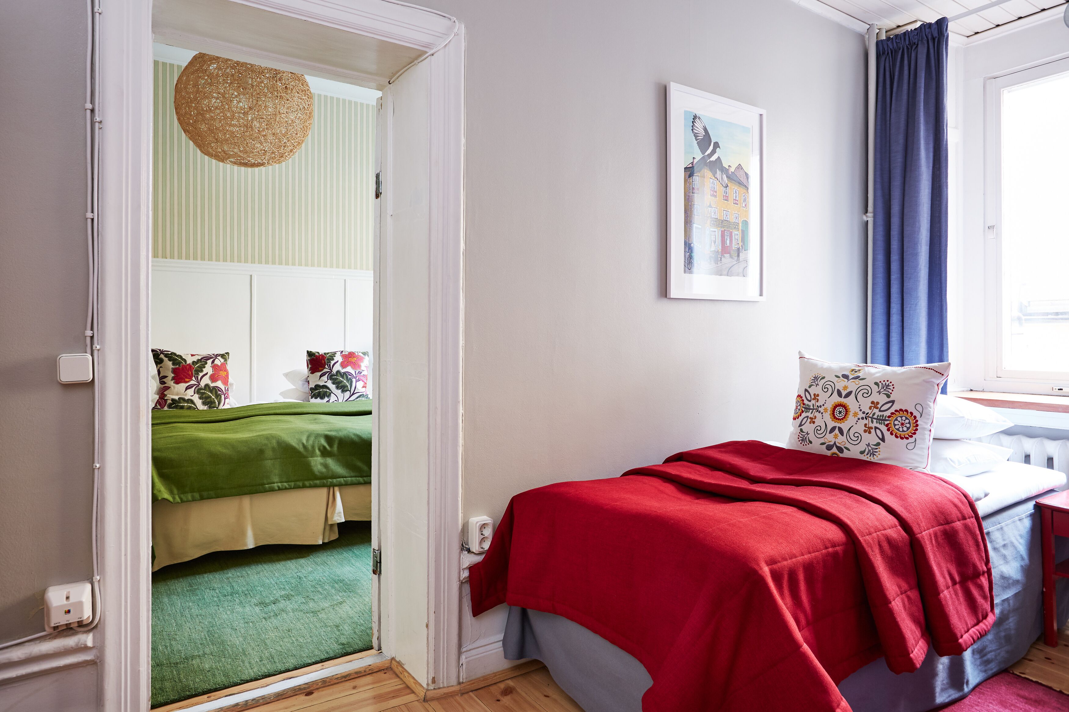A bedroom with a bed, a window with curtains, a door leading to another room, and a wall with a framed picture.