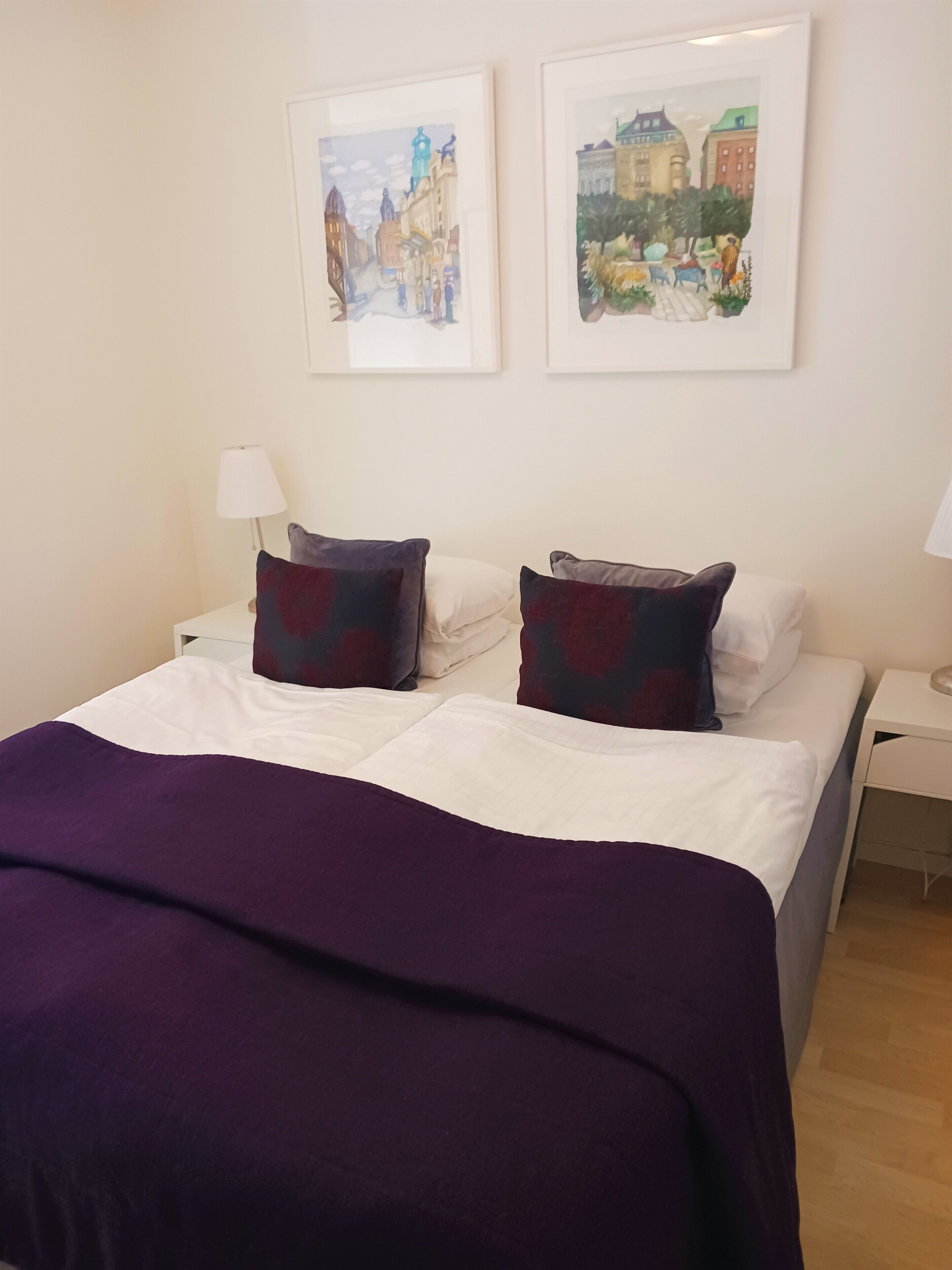 Ein ordentlich bezogenes Bett mit weißen Leinentextilien und einer violetten Tagesdecke, umrahmt von zwei Kissen in verschiedenen Dunkel- und Helltönen. Darüber an der Wand hängen zwei gerahmte Kunstwerke mit Stadtansichten.