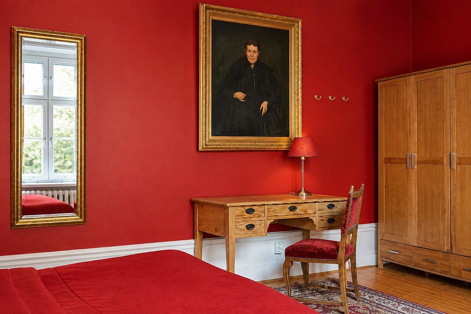 Une pièce avec un mur rouge, un bureau en bois doté de tiroirs, une chaise, un meuble et un portrait encadré.
