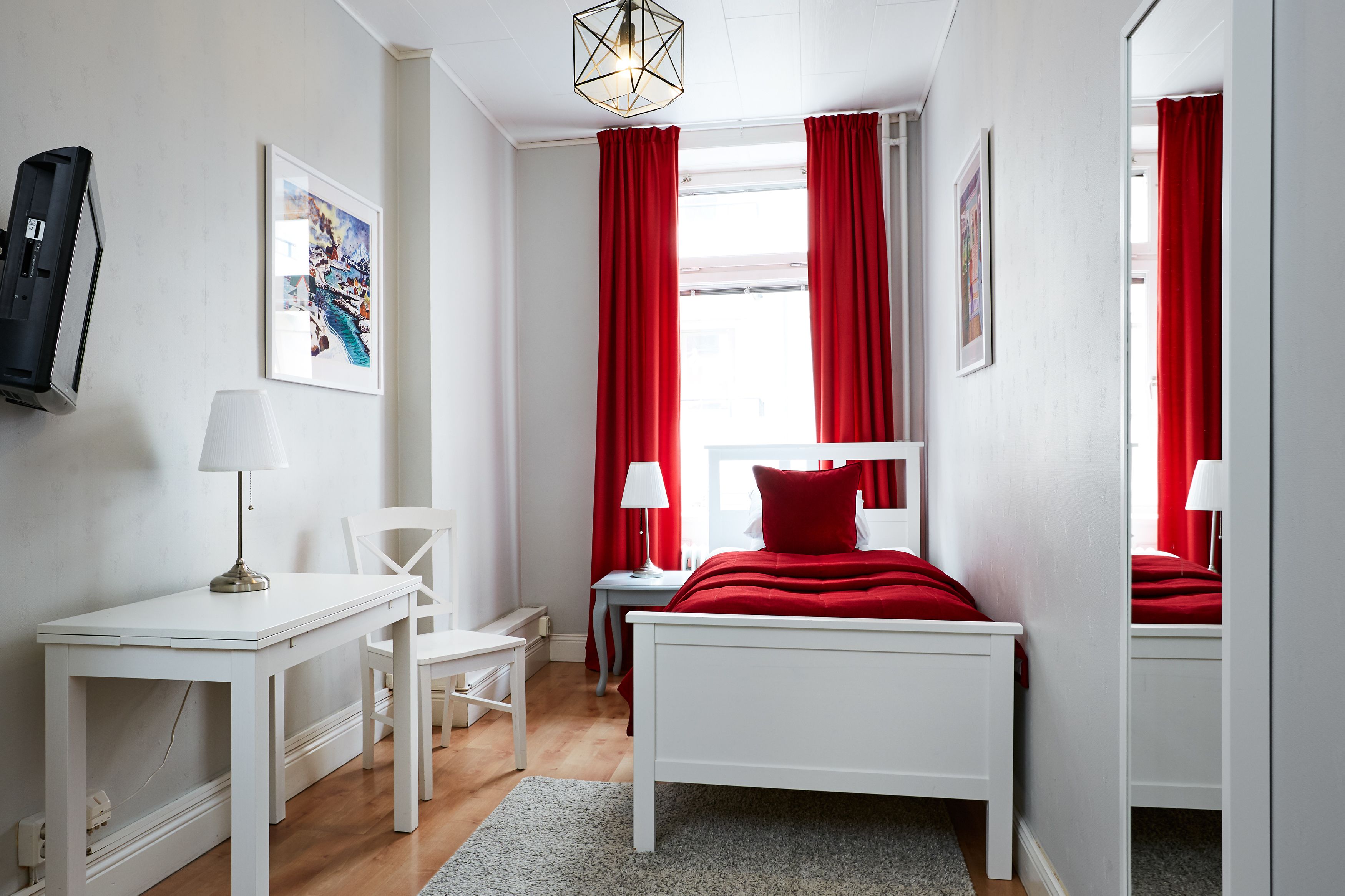 Et soveværelse med en hvid seng, røde gardiner, et skrivebord med lampe og en stol.