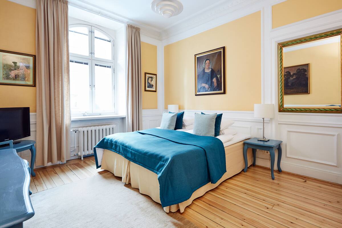 Una camera da letto con un letto, un comodino, una televisione, una finestra con tende e quadri appesi alle pareti.