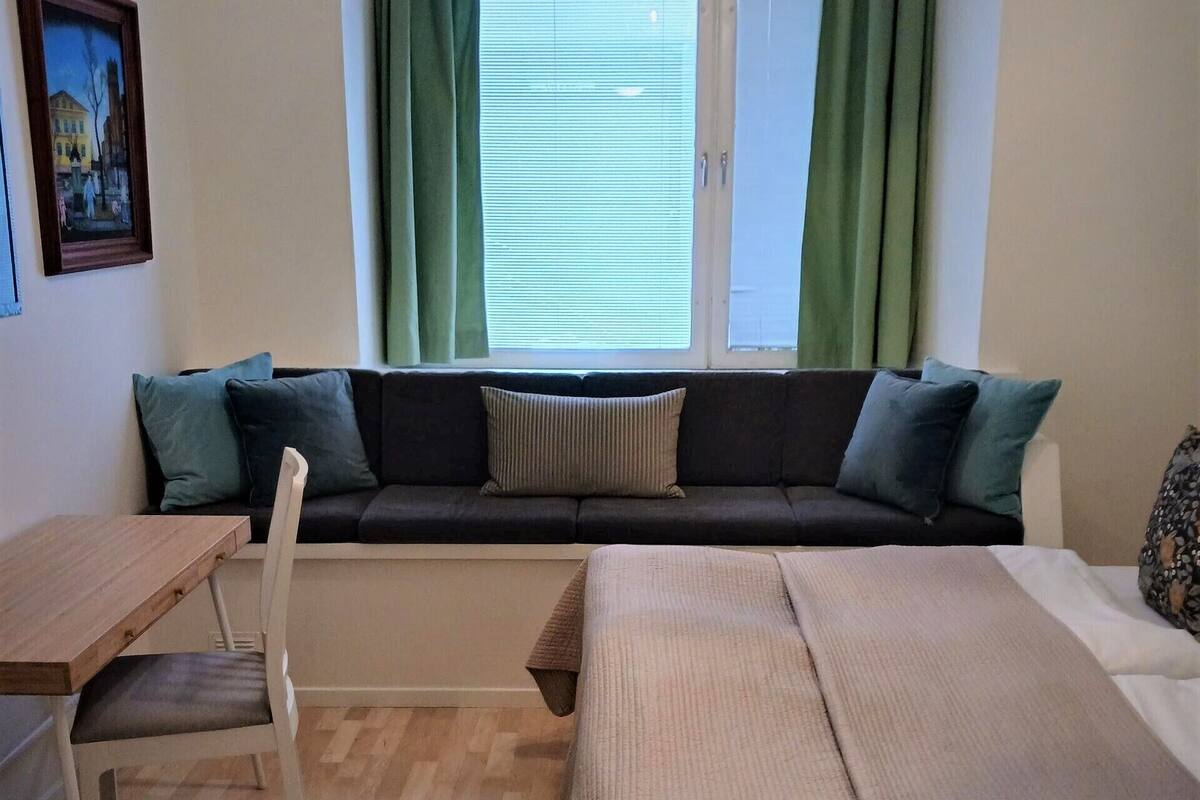 Una stanza accogliente con un letto, un tavolino e una panca. Si vede una finestra con tende, un quadro al muro e un lampadario.