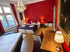 A hotel room with a bed, a wooden table, a lamp, a radiator, and a large painting on the wall.