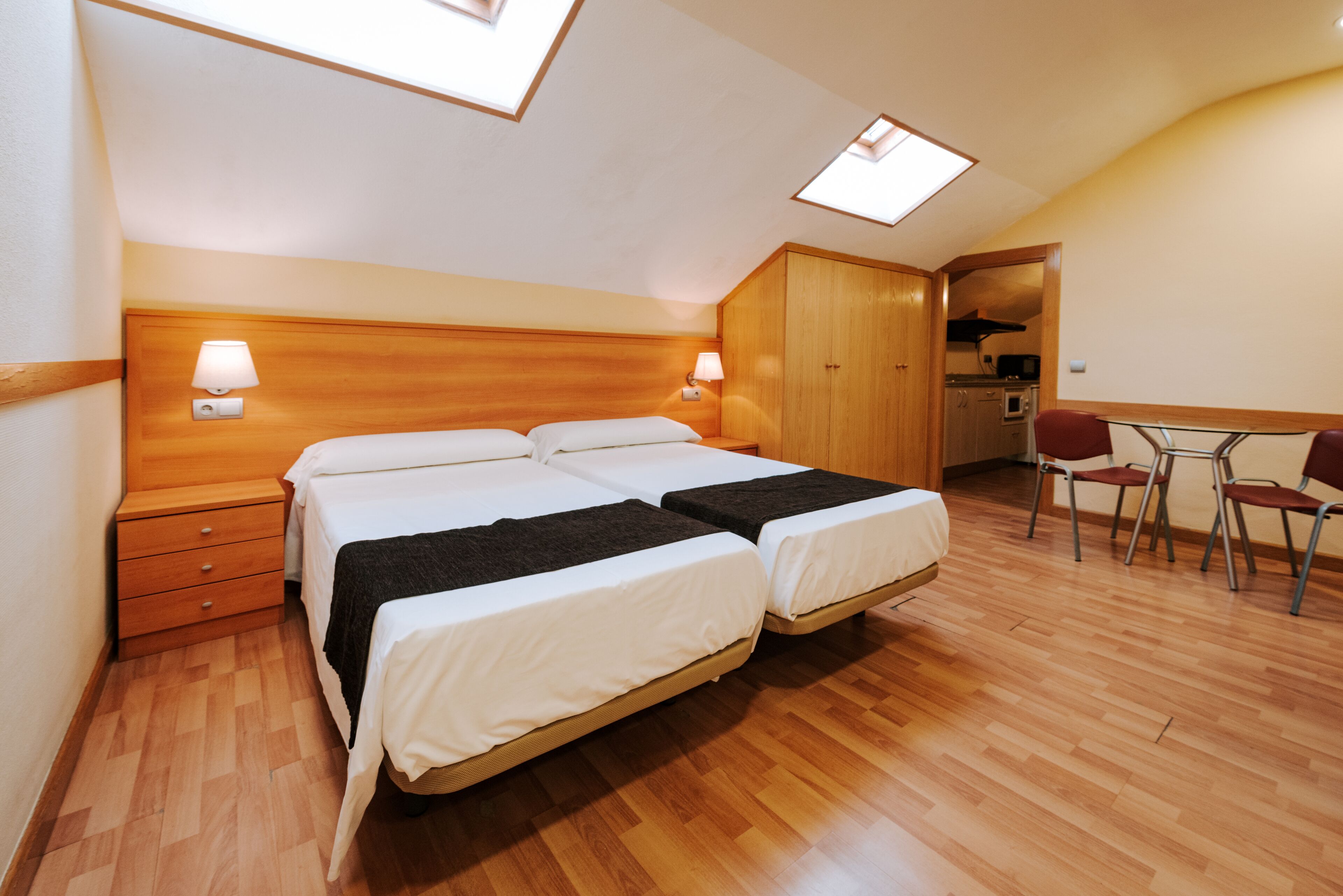 Un dormitorio con dos camas, cabeceros de madera, lámparas de noche, un escritorio con sillas y una kitchenette.