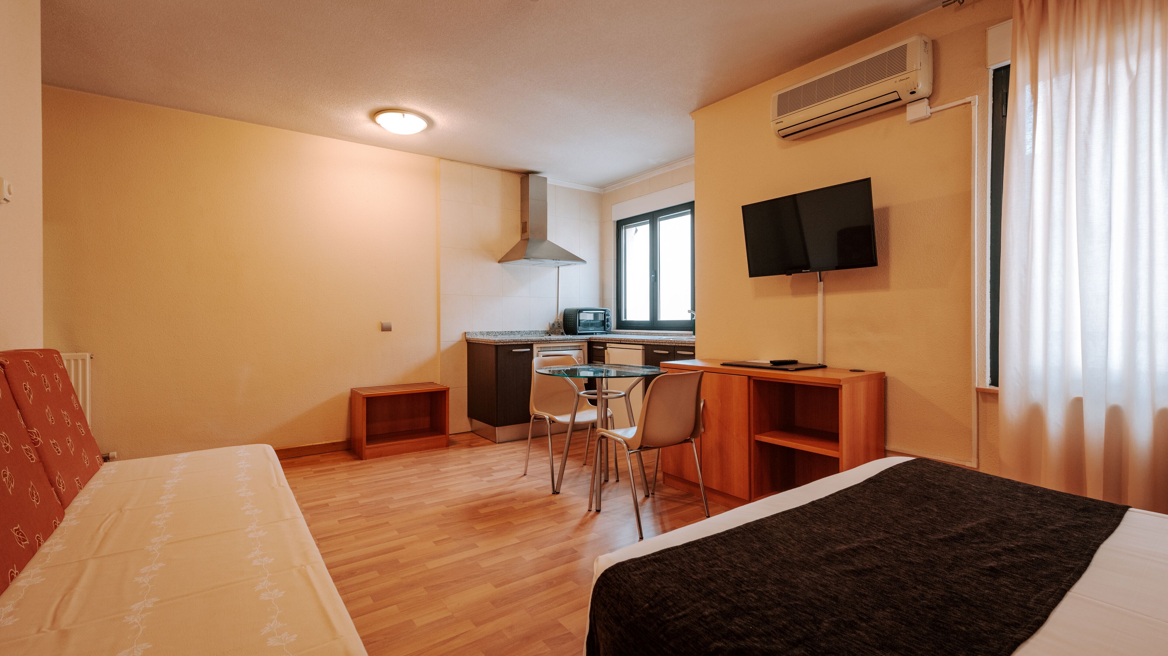 A compact hotel room with a bed, a small kitchenette, a dining area with a table and chairs, a TV mounted on the wall, and a window with curtains.