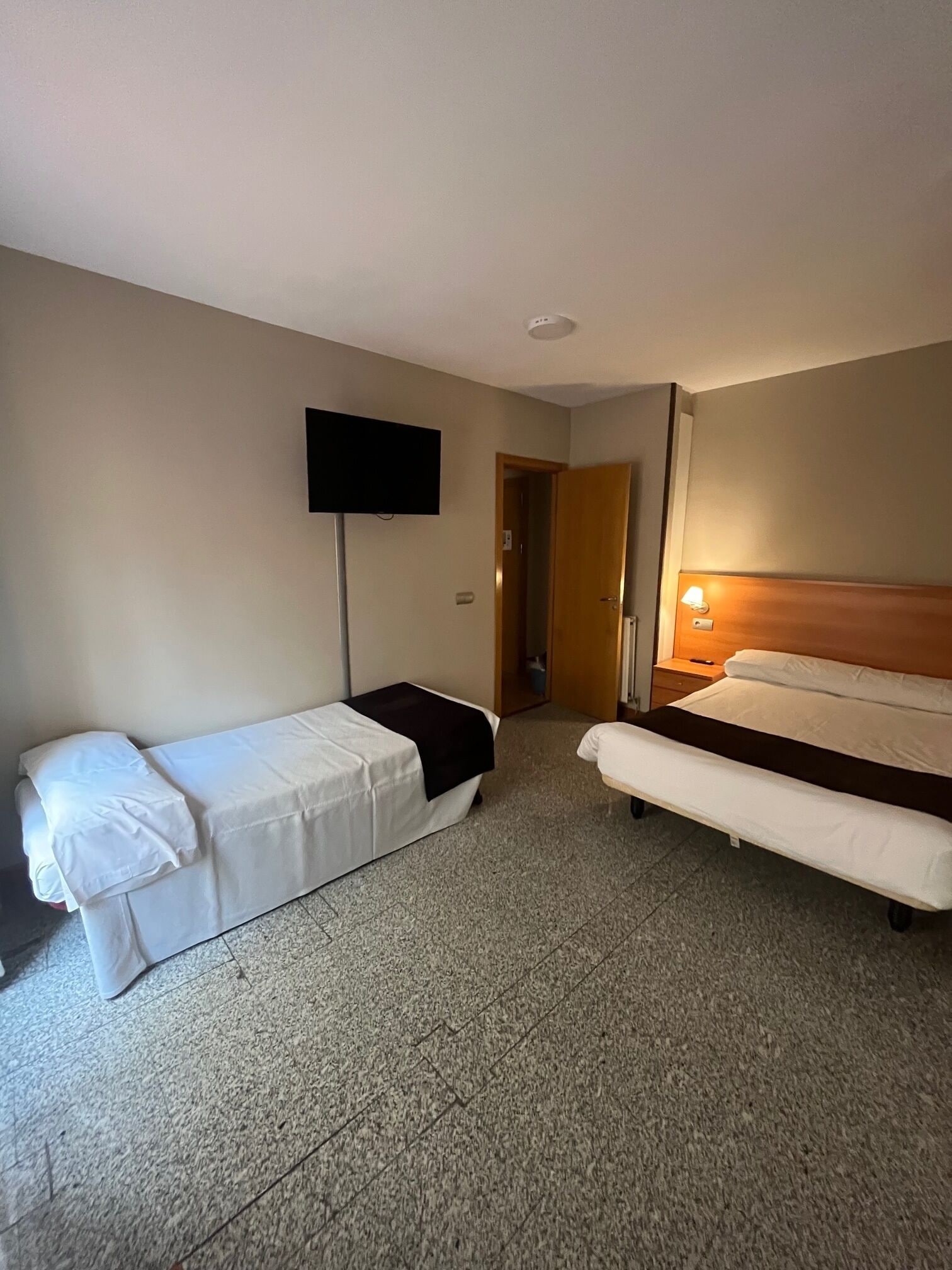 A hotel room with two beds, a flat-screen TV mounted on the wall, and a bedside table with a lamp.