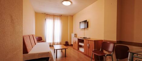 Ein kleines Hotelzimmer mit Bett, Esstisch, Schreibtisch, Fernseher und einem Fenster mit Vorhängen.