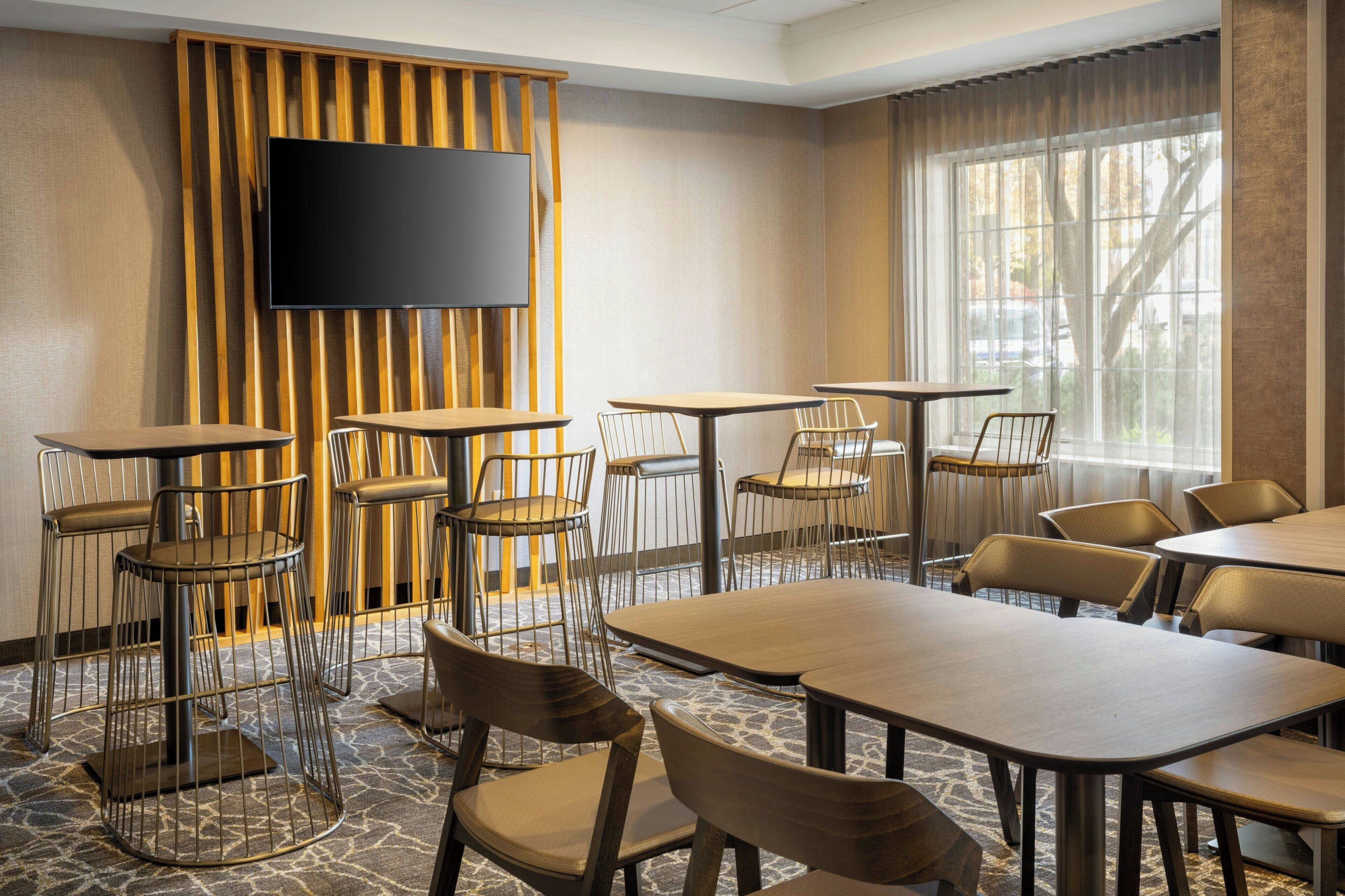 A conference room with a large flat-screen TV mounted on a wooden panel wall, surrounded by multiple tables and chairs.