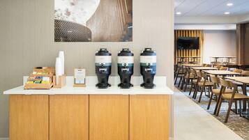 A hotel reception area with a counter, three coffee dispensers, a television, and a seating area with tables and chairs.