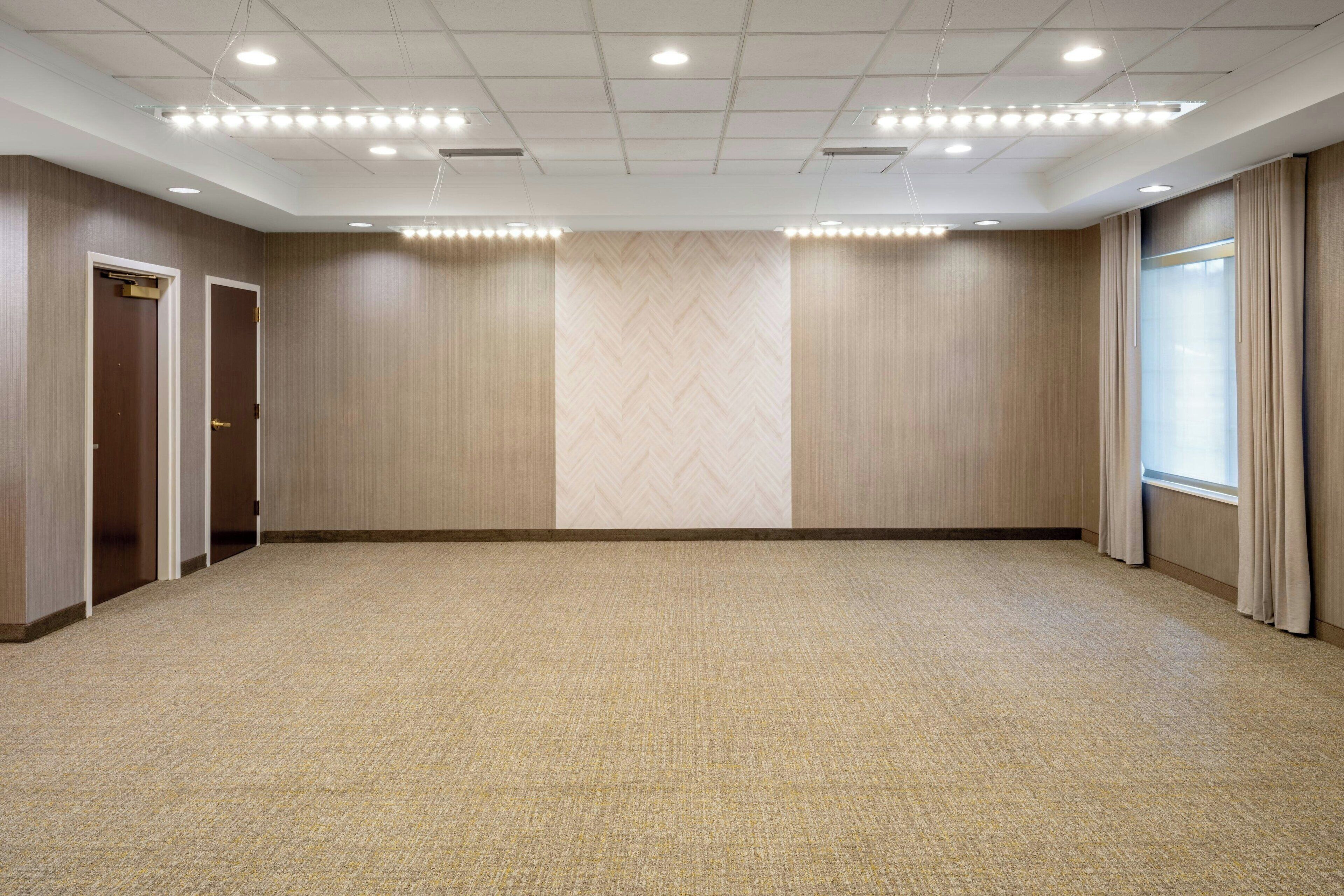A large, empty conference room with a carpeted floor, wooden panel walls, and a large window with curtains.