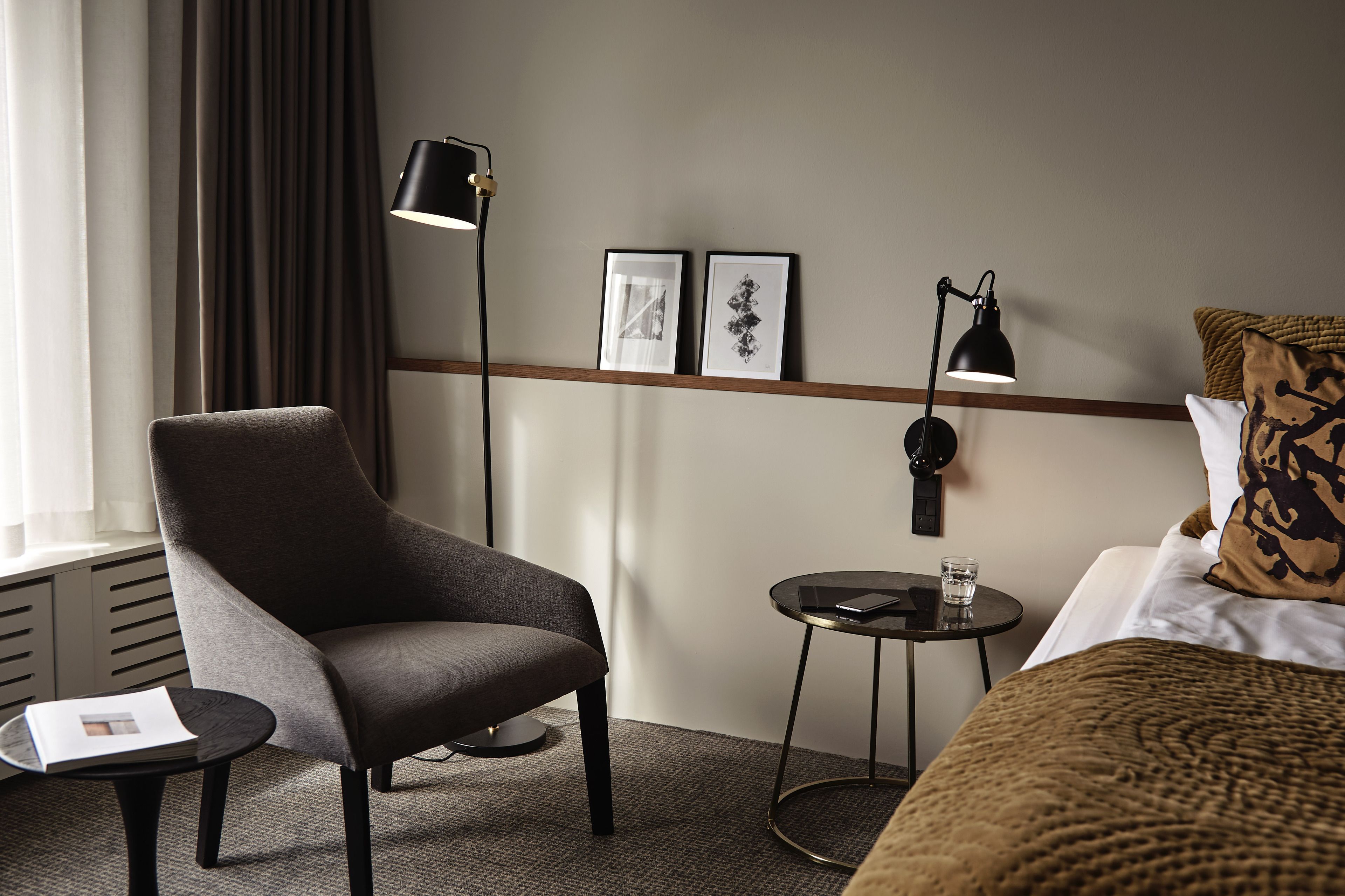 Ein Hotelzimmer mit grauem Sessel, einem kleinen runden Tisch mit einem Glas, einem Bett mit brauner Decke und zwei Wandlampen.