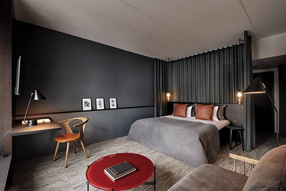 Moderni hotellihuone, jossa on sänky, työpöytä, tuoli ja punainen pöytä.