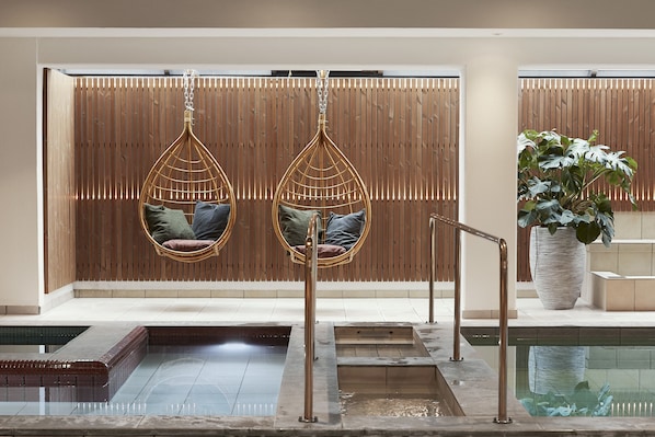 Un área de spa con dos sillones colgantes, una pared de madera, una planta en maceta y una piscina.
