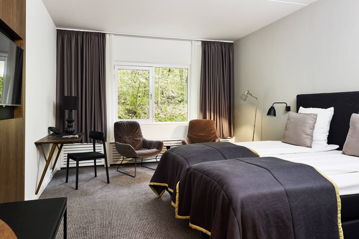 Hotellihuone, jossa on sänky, kaksi tuolia, työpöytä lampun kanssa, televisio ja ikkuna, jossa on verhot.