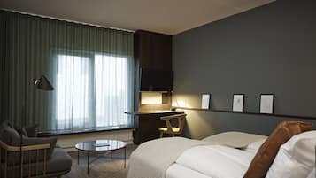 A modern hotel room with a sofa, a small round table, a desk with a lamp, and a large window with curtains.