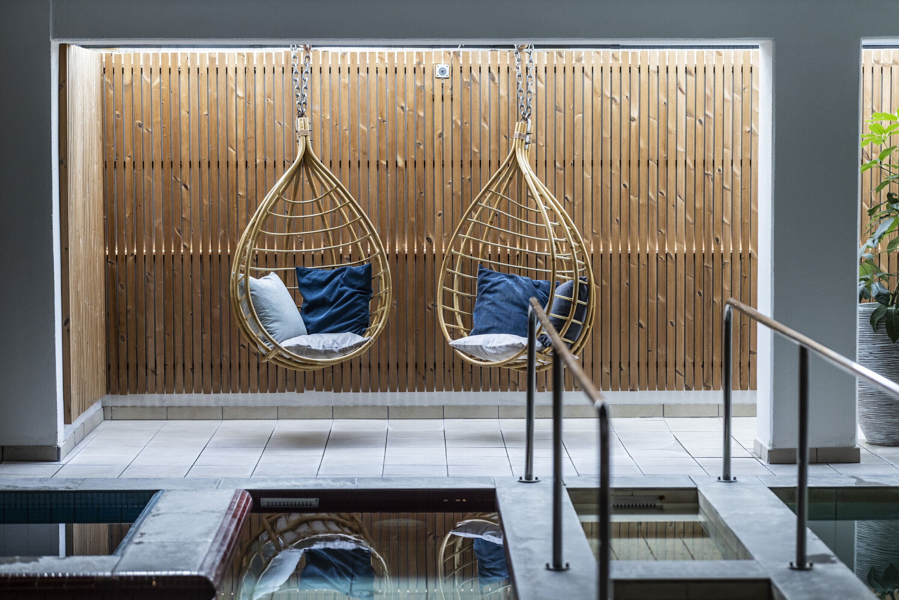 A modern spa area with two hanging chairs, a wooden wall, and a pool.
