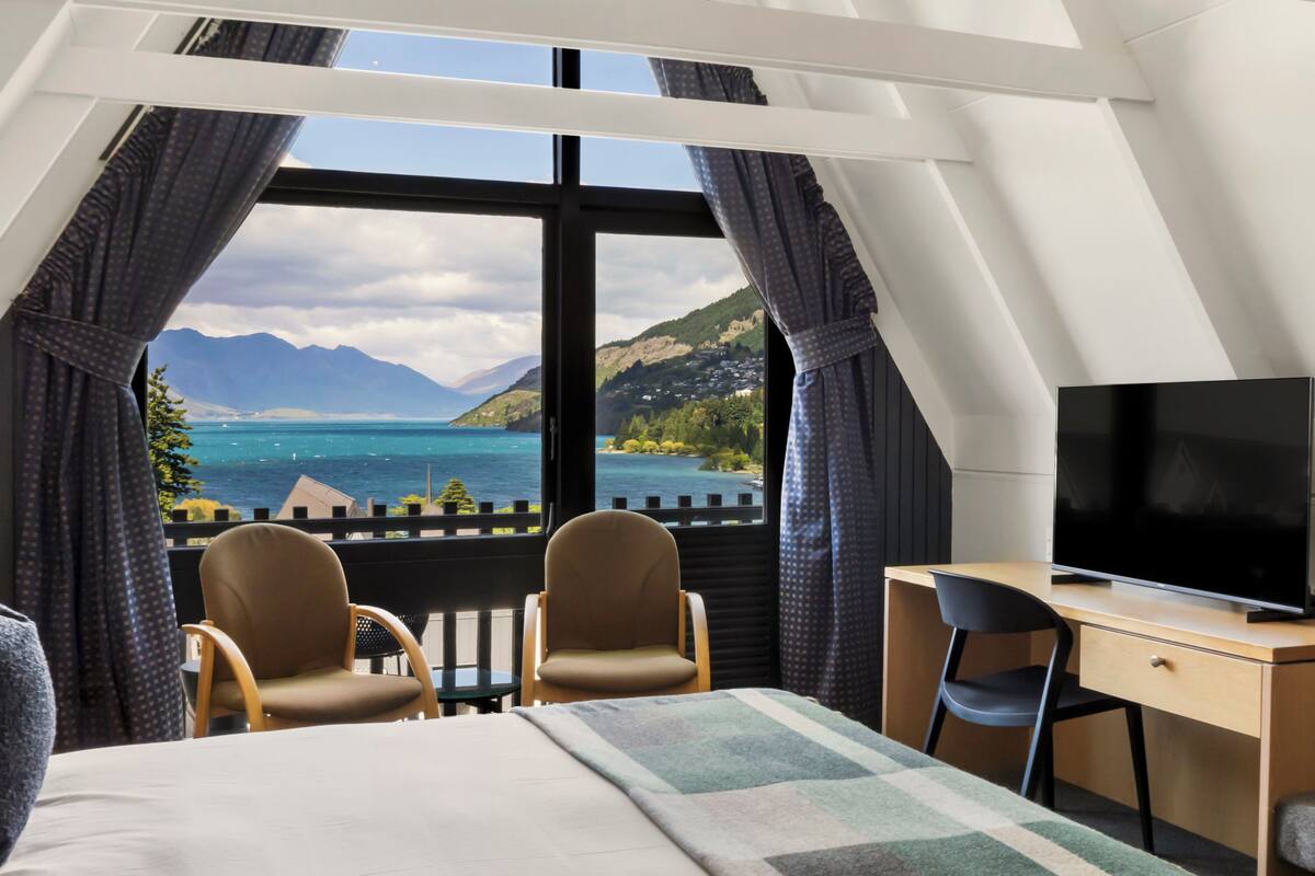 A hotel room with a bed, two chairs, a desk with a TV, and a large window offering a view of mountains and a lake.