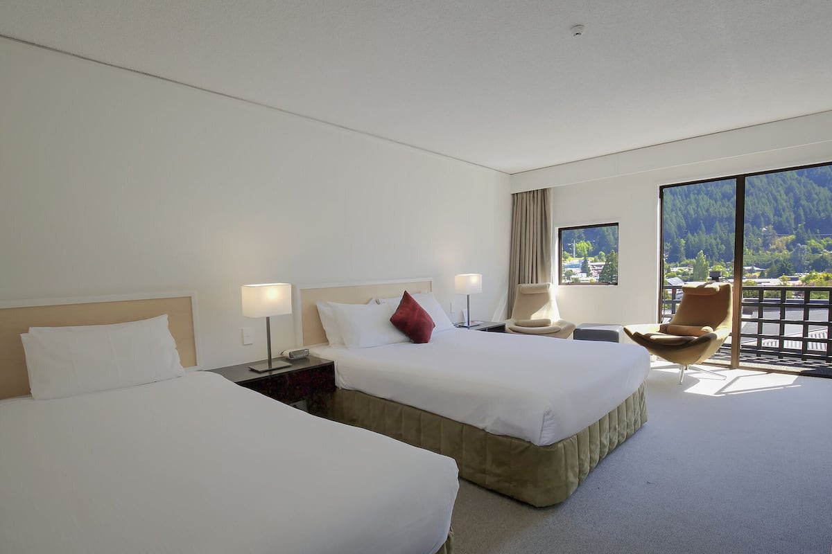 A hotel room with two beds, a desk, a chair, and a large window offering a view of a mountainous landscape.