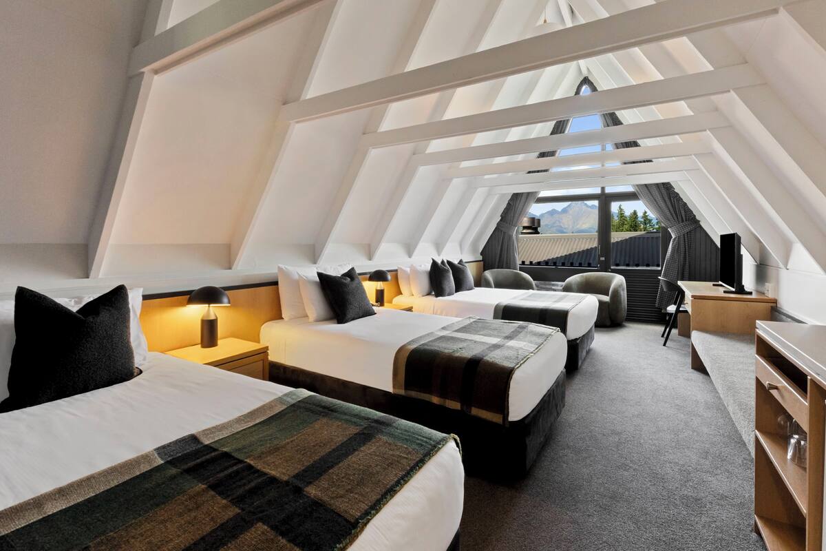 A modern hotel room with two beds, a sofa, a desk, and a view of mountains through large windows.