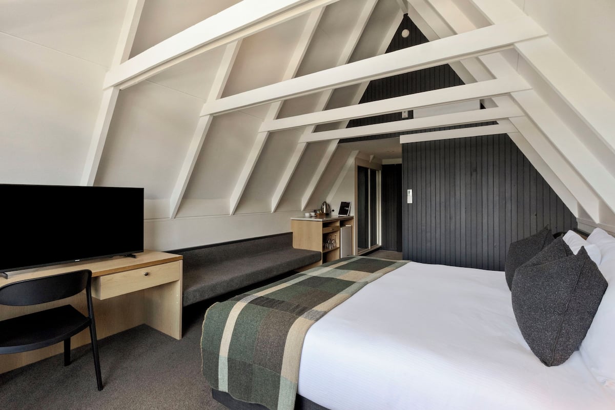 A modern hotel room with a sloped ceiling, a bed, a desk, a chair, and a television.