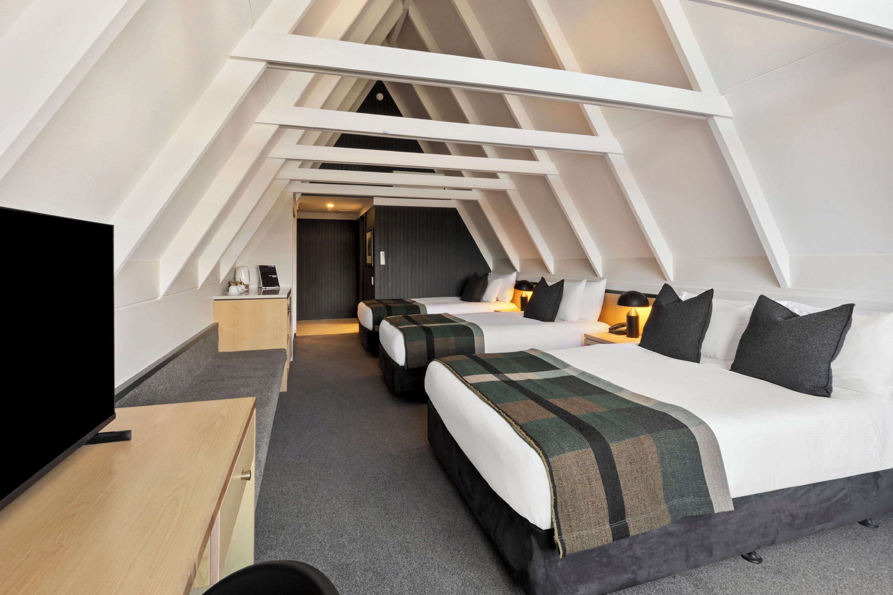 A modern hotel room with a sloped ceiling, a large bed, two smaller beds, a desk, and a television.