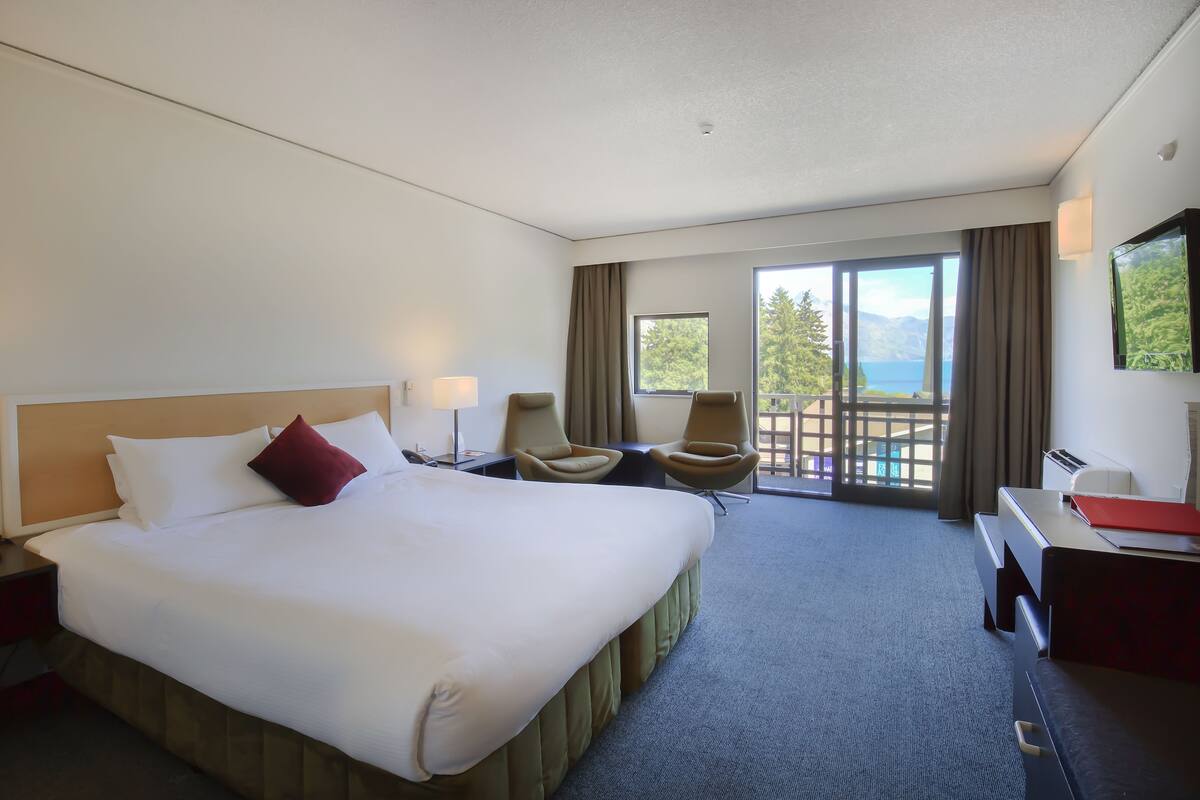 A hotel room with a large bed, two armchairs, a desk, a TV, and a balcony with a view of the mountains and lake.