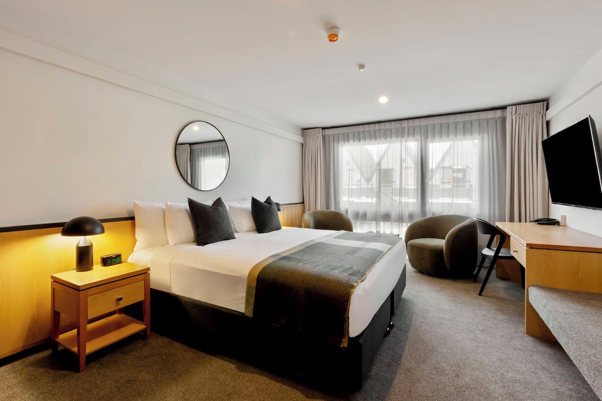 A hotel room with a bed, bedside table, lamp, chair, desk, TV, and a large window with curtains.