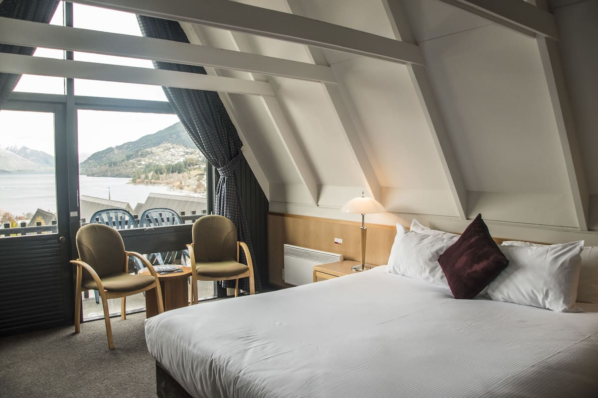 A hotel room with a bed, two chairs, a table, a lamp, and a view of a lake and mountains.