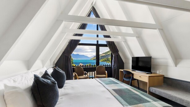 Une chambre moderne de forme triangulaire, équipée d’un lit, d’un bureau, d’une télévision et offrant une vue sur un lac et des montagnes.