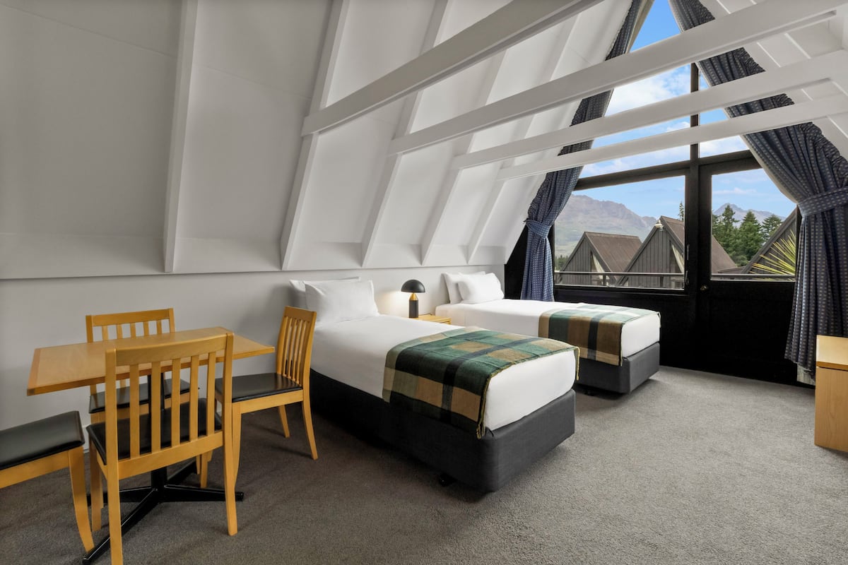A hotel room with a high ceiling, a wooden table, two beds, and a view of mountains through large windows.