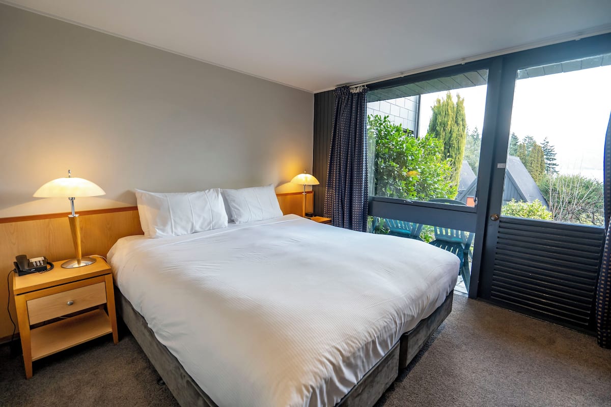 A hotel room with a large bed, two bedside lamps, a nightstand with a phone, and a window with a view of greenery and buildings.