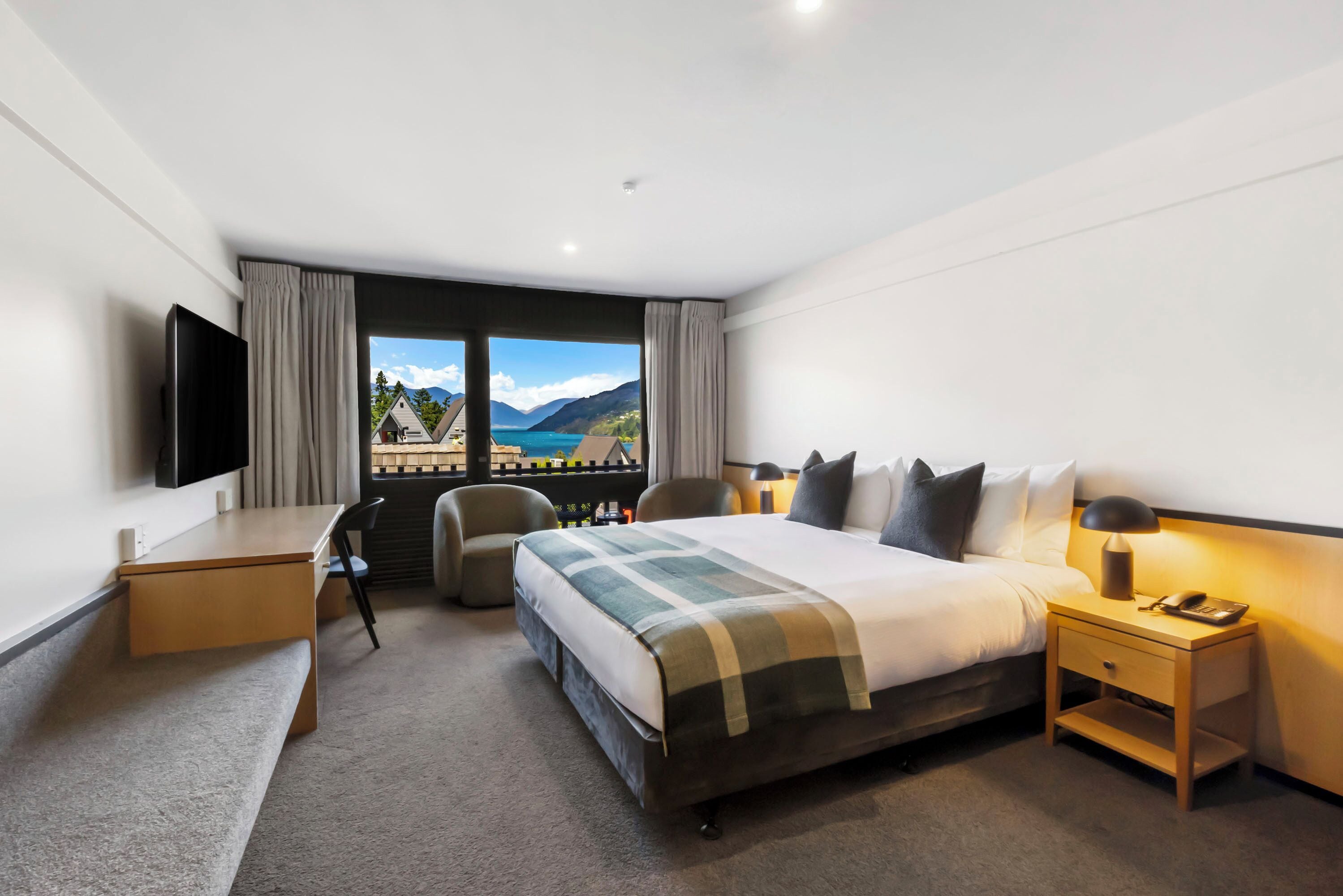 A hotel room with a bed, a desk, a chair, a TV, a nightstand, and a view of mountains and a lake.