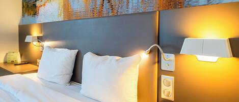 A hotel room with a large bed, two white pillows, a nightstand with a lamp, and a wall-mounted light fixture.