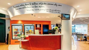A modern hotel reception area with a curved red counter, a welcome sign, and a view of the lobby. - B&B HOME Rueil-Malmaison Gare (Rueil-Malmaison)