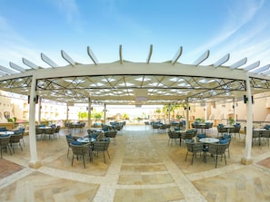 Un espace de restauration en plein air doté d’une terrasse couverte, de plusieurs tables et chaises, et offrant une vue sur les bâtiments en arrière-plan.