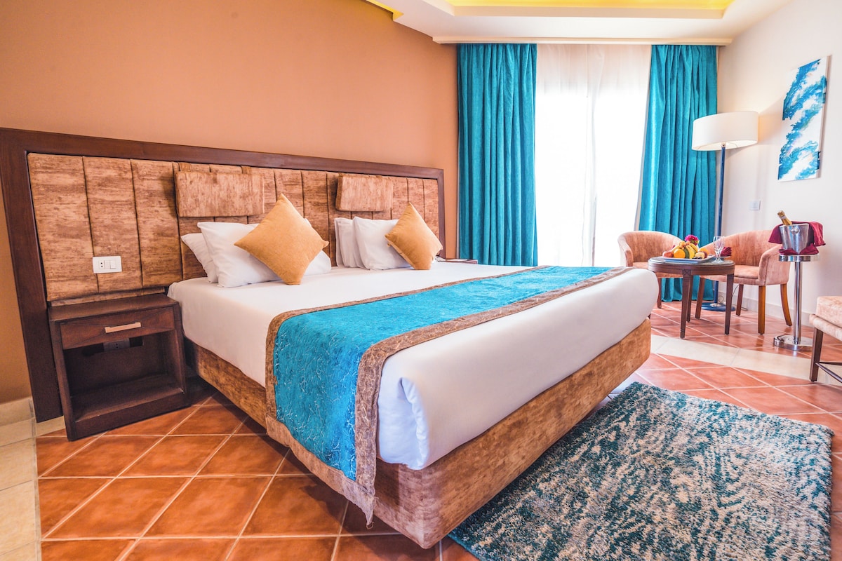 A hotel room with a large bed, a wooden headboard, a nightstand, a desk with a lamp, and a view of the outside through a window with blue curtains.