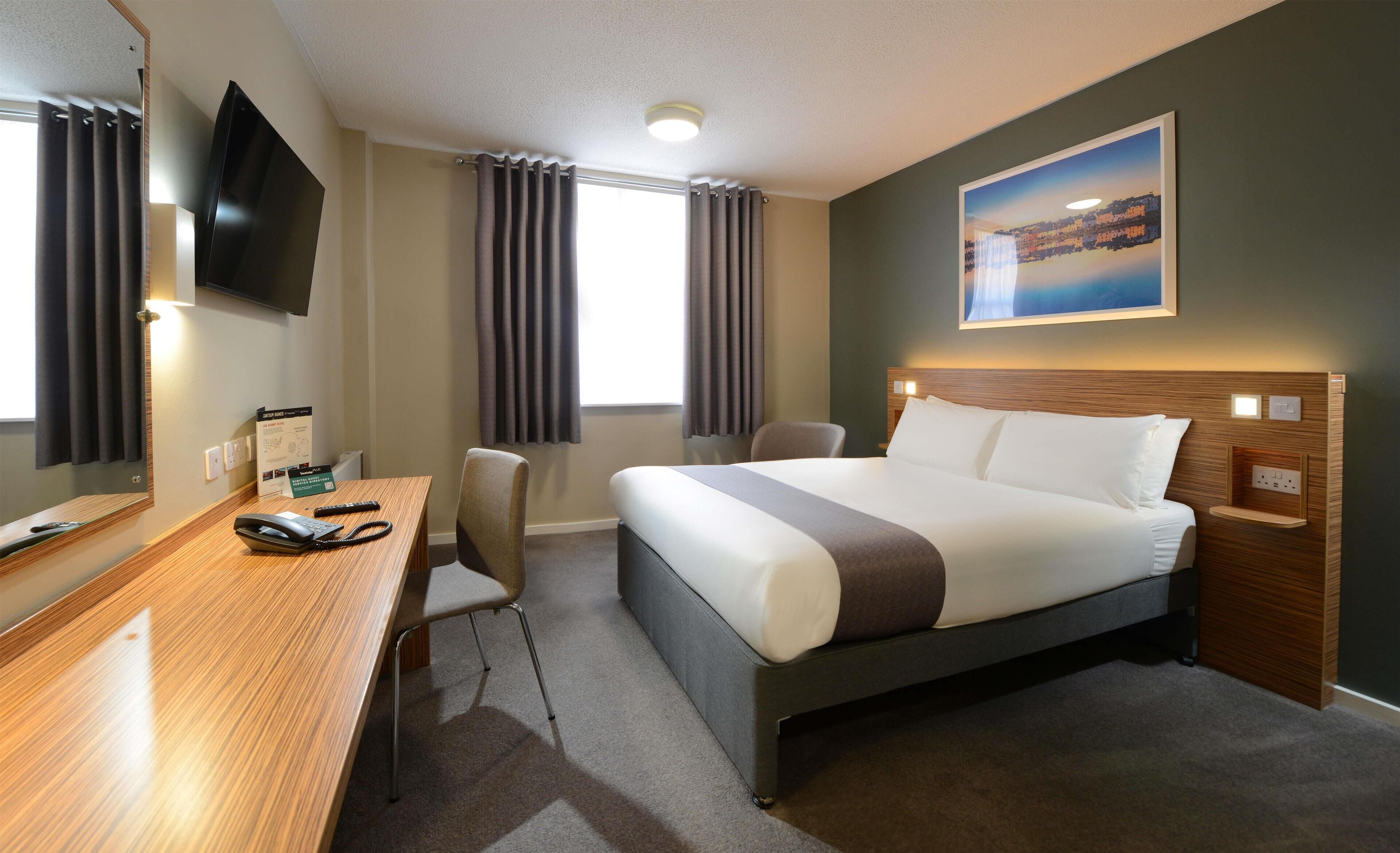 A hotel room with a bed, a desk, a chair, a TV, and a painting on the wall.